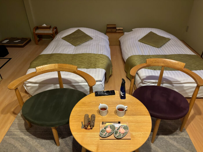 洋室ツインの客室内。ロータイプのベッドが2つ並んでいる。ベッドの足元には椅子が2脚と丸テーブルが1つ。テーブルの上にはリモコン、ルームキー、消毒スプレーがある。ウェルカムドリンクの紅茶、りんご大福が2つずつある。