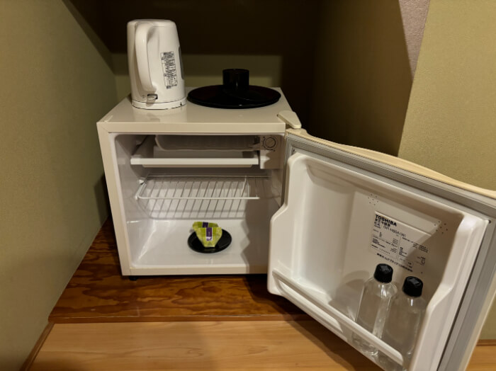 ミニ冷蔵庫。冷蔵庫の上にはポットと茶筒がある。冷蔵庫の中にはペットボトル2本と黒豆ゼリーが2つ入っている。